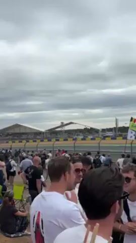 Here is a description of the video:

The video shows a crowd of people gathered near a racetrack, with cars speeding past on the track. The sky is cloudy, and there are tents and buildings visible in the background. The people in the foreground are standing around talking, and they appear to be enjoying the event. Several flags are visible at the event, including one for Belgium and a green and white flag with the words "100 years". The video seems to have been taken by someone within the crowd.
