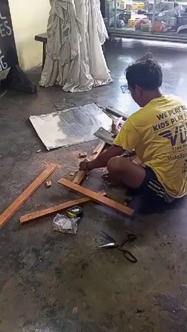 A person wearing a yellow t-shirt and dark shorts is sitting on the floor and using a saw to cut a wooden plank. There are several other wooden planks scattered around the person on the floor, along with a roll of tape and a pair of scissors. A large, reflective sheet of metal is also on the floor, partially underneath the planks. In the background, there is a table with some draped fabric and a display case. The floor appears to be concrete.