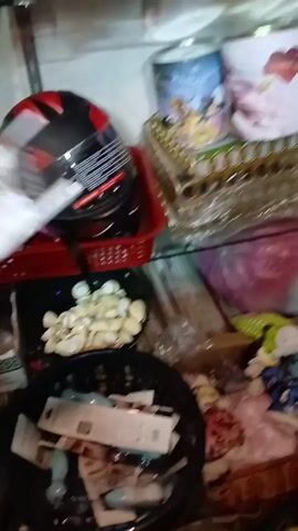 The video shows a woman in a shop. She is wearing a headscarf and is holding a small electric coffee whisk in its packaging. There are shelves behind her, with a helmet and various other items. She looks at the camera and speaks.