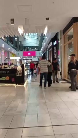 The video shows the interior of a shopping mall. The camera moves along a tiled walkway, capturing several individuals walking in various directions. People include men and women of diverse ages, some carrying shopping bags. Shops line both sides of the walkway, with displays visible through the windows. A kiosk with food products is on the left. The mall is multi-storied, with a view of the upper levels and a glass ceiling allowing natural light. Lighting fixtures are visible. The environment seems populated and bustling.
