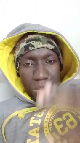 A close-up, eye-level shot shows a man wearing a grey hoodie with a yellow lining and drawstrings, and a camouflage knit hat. He is looking directly at the camera with a slightly serious expression, making a gesture with his hand in front of his face, specifically a pinching motion with his fingers. The background is a plain white wall.
