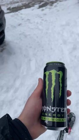 The video begins with a close-up shot of a hand holding a black Monster Energy Zero Sugar can against a backdrop of snow and skis. The camera then pans upwards to reveal a black Ram truck parked in a snowy environment. The truck is customized with Monster Energy branding, including green claw marks and the Monster logo on the hood and grill. The environment shows falling snow and other vehicles parked nearby, suggesting a winter outdoor setting, possibly a ski resort or parking lot.