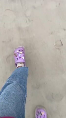 The video begins with a close-up shot of sandy ground with footprints and then pans up to reveal a beach scene. A person wearing purple crocs and blue jeans is visible at the bottom of the frame. The camera then focuses on the ocean, showing gentle waves rolling onto the sandy shore under a cloudy sky. The horizon is visible with a hint of land in the distance.