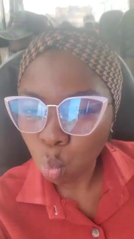 A young woman with dark skin, wearing a red shirt and a patterned headband, is taking a selfie on a bus. She is wearing large, light pink cat-eye glasses with blue lenses. In the background, another person wearing a dark cap is visible. The woman puckers her lips for the camera, then smiles before puckering again. The video captures her expressions and the interior of the bus.