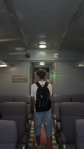 A person wearing a white t-shirt, denim shorts, and a black backpack walks through a doorway at the end of a train car. The train car has rows of dark gray seats on either side, and the walls and ceiling are a metallic gray. The doorway leads to another section of the train, visible as dimly lit. A sign is mounted on the left wall near the doorway. The lighting in the car is from overhead circular fixtures.