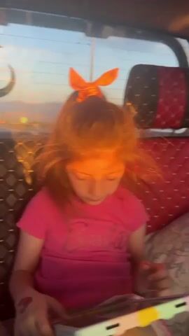 A young girl with red hair and an orange bow is sitting in the back of a vehicle, looking down at a tablet she is holding. She is wearing a pink "Barbie" t-shirt. The vehicle's interior is visible, showing a red and black diamond-patterned seat. The back window offers a glimpse of the outside environment, where a setting sun is visible. The girl appears absorbed in the tablet, her fingers moving across the screen.