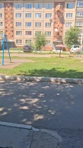 The video pans across a courtyard in front of a multi-story apartment building. The building appears to be made of concrete and has many windows. There are satellite dishes attached to the side of the building. In the courtyard, there is a playground with swings, a seesaw, and benches. There is also a grassy area and trees. Several cars are parked in front of the building. The sky is blue and the sun is shining.