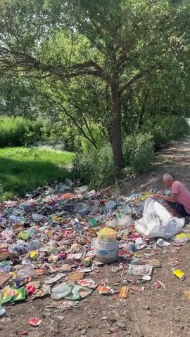 The video shows a scene of severe littering alongside a dirt path, adjacent to a grassy area leading down to a body of water. The ground is almost entirely covered with various types of trash, including plastic bottles, food wrappers, cans, and paper debris. An elderly person is squatting next to a large pile of trash, sorting through the litter and placing items into a white plastic bag. There's also a white bucket with a yellow lid near the center of the scene.  A large tree provides shade over part of the area, and the background features lush greenery.