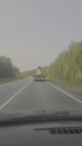 The video shows a view from inside a car looking forward at a highway. The road is asphalt with white lane markings. On the right side of the road is a dense forest of green trees. The sky is overcast and hazy. In the distance, centered in the road, a large yellow object is being transported on the back of a flatbed truck. The object appears to be the fuselage of a helicopter, with its cockpit and main body visible. The truck is traveling away from the camera.