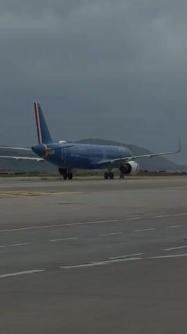 A blue ITA Airways plane is on an airport tarmac. The plane is facing away from the viewer and the Italian flag is on the tail. Mountains can be seen in the distance and the sky is overcast.