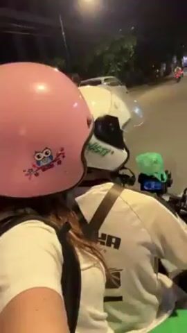 Two people are riding on a motorcycle at night. The person in the back is wearing a white shirt and a pink helmet with an owl sticker. The person driving is wearing a white helmet and a white shirt with black Puma lettering. The street is lit by streetlights and Christmas lights, and other vehicles can be seen in the distance.