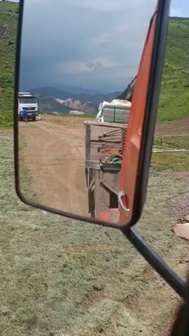 The video is filmed from the perspective of someone looking into a side mirror of a vehicle. The mirror reflects a dusty, rural landscape. In the mirror, a white truck is visible on a dirt road, followed by another truck, likely the one from which the video is being filmed, which is loaded with what appears to be building materials. The background shows rolling hills with green vegetation and distant mountains under a cloudy sky. Outside the mirror, the foreground is dominated by dry, grassy ground and the side of the vehicle's orange side mirror housing.