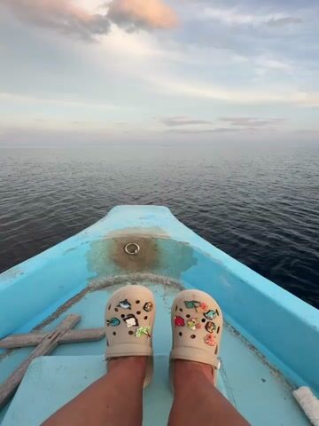 The video presents a first-person perspective looking forward from the bow of a small boat. A person's bare feet, clad in light beige Crocs adorned with numerous Jibbitz charms, rest on the light blue floor of the boat. The charms depict a variety of sea-themed elements (fish, coral, palm trees) and food items (tacos, pizza).

The boat is a simple, light blue vessel, showing signs of wear and rust near a metal ring on its floor at the bow. A light-colored wooden plank lies on the boat floor to the left.

Beyond the boat, a vast, calm expanse of dark blue ocean stretches to the horizon with gentle ripples on its surface. The sky features soft pastels of pale blue, white, and faint pink/orange, characteristic of sunrise or sunset, with wispy clouds scattered across the horizon.

The boat appears to be moving slowly and steadily across the tranquil waters. The scene evokes a sense of peace and relaxation, likely depicting a leisurely boat ride or transfer within the serene environment of Ukulhas, Maldives, given the location context. No visible text is present in the video.