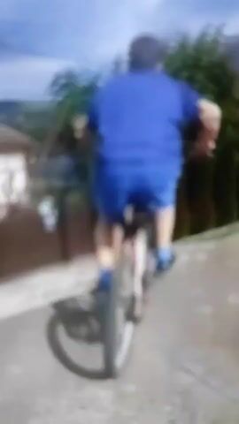 A person wearing a blue t-shirt and blue shorts is riding a bicycle. The person is wearing a hat and glasses. The person is riding on a paved path. There are trees in the background. The sky is cloudy. The video is blurry.