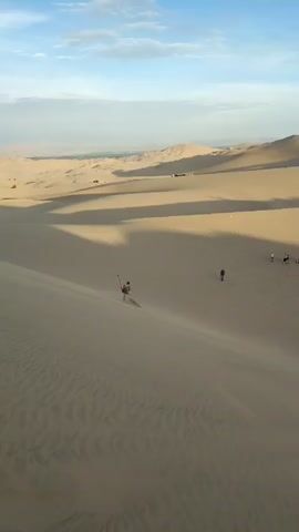 The video shows a man sand skiing down a dune in a desert landscape. Several other people are standing in the background, near what appear to be sandboards, suggesting they may be engaging in sandboarding activities. The desert environment is characterized by rolling sand dunes under a partly cloudy sky. The camera captures the scene from an elevated perspective, emphasizing the vastness of the desert and the steepness of the dune the man is skiing down.