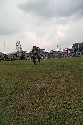 Here is a concise description of the video:

The video shows a person riding a bicycle across a grassy field. A crowd of people is visible in the background, along with some tents and structures. The sky is cloudy. The video is taken from a slightly angled perspective.