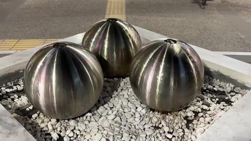 The video captures three large, spherical, metallic fountains operating in an urban setting during the evening or night. Each sphere, appearing to be made of polished stainless steel, features a ribbed design that reflects the surrounding light, creating shimmering visual effects. Water continuously bubbles from the top center of each sphere, flowing gracefully down its surface and into a collection area filled with white decorative pebbles. The spheres are housed within a square planter with light-colored, possibly marble or concrete, edges.

In the background, a paved pedestrian area is visible, complete with yellow tactile paving strips. Partially obscured by the low light, distant figures of people and bicycles can be seen moving. The primary focus is on the tranquil water feature, with the constant, soothing sound of flowing water clearly audible. The scene is static, offering a peaceful and aesthetically pleasing view of the illuminated fountains. No visible text is present in the video frame. The location is Karditsa, Greece.