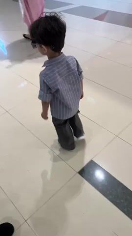 A young child wearing sunglasses, a striped collared shirt, and dark pants walks across a tiled floor. Another young child in a white shirt and dark pants walks towards the first child. A person in a pink outfit stands in the background. The environment appears to be an indoor public space with polished tile flooring.