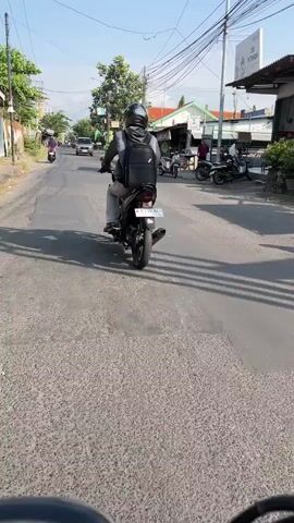 The video shows a street scene in Sidoarjo, East Java. A person is riding a scooter down a paved road. The sun is shining, indicating it is daytime, and the weather appears to be clear.

The road is lined with buildings and trees. There are other scooters and motorbikes on the road, as well as some pedestrians. In the background, there is a sign for "ZHE PETSHOP". The overall atmosphere is one of everyday life and activity in a semi-urban environment. The time of day appears to be late morning or early afternoon, given the bright sunlight and the long shadows. The scene is bustling with traffic, suggesting a moderately busy area. The architecture of the buildings and the type of vehicles suggest a Southeast Asian setting, which is confirmed by the location context of Sidoarjo, East Java. The rider in the foreground is wearing a helmet and a backpack, indicating safety precautions and preparedness for travel. The presence of a pet shop further suggests a residential or mixed-use area.
