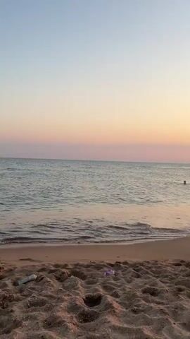 The video opens with a view of a beach at sunset. The sand is in the foreground, with the ocean stretching out to the horizon. The sky is a gradient of soft pinks and oranges. A person is visible swimming in the ocean in the distance. A small boat appears briefly further out at sea. Gentle waves lap against the shore. The lighting suggests it is late evening.