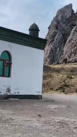 THE VIDEO SHOWS A SIDE VIEW OF A SMALL, WHITE BUILDING WITH A DARK GREEN ROOF AND TRIM. A SINGLE ARCHED WINDOW WITH A BROWN FRAME AND GREEN GLASS IS VISIBLE ON THE SIDE OF THE BUILDING. THE BUILDING HAS A SMALL, DOME-LIKE STRUCTURE ON ITS ROOF. TO THE RIGHT OF THE BUILDING, A STEEP, ROCKY MOUNTAIN SLOPE IS VISIBLE. THE GROUND IN FRONT OF THE BUILDING IS DIRT AND GRAVEL. THE SKY IS OVERCAST. A PERSON IS VISIBLE IN THE BACKGROUND ON THE MOUNTAIN SLOPE.