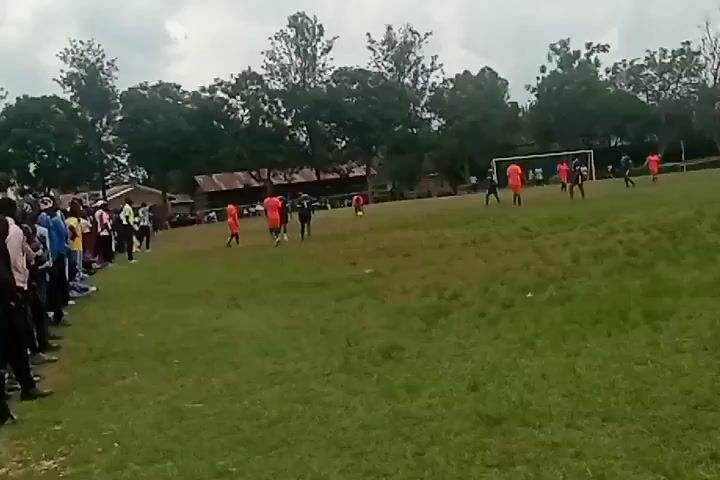 The video shows a soccer game in progress on a grassy field. There are two teams playing, one in orange jerseys and the other in black. There is a small crowd of people standing along the sideline, watching the game. There are trees in the background, and the sky is overcast.