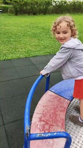 A young child with curly blonde hair and wearing a striped jacket and red shorts is on a merry-go-round in a park. The child is holding onto the blue railing and smiling at the camera. The merry-go-round is made of blue metal and has a metal platform with a diamond pattern. The background shows a grassy area with trees and a building. The camera then pans to show the merry-go-round spinning, with the child visible for a moment before disappearing out of frame. The video continues to show the empty merry-go-round as it spins. The surroundings of the park are visible, including green grass and trees. The weather appears to be overcast.