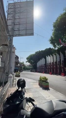The video pans across a street scene under a bright sun. In the foreground, the seat and handlebars of a parked motorcycle are visible. A black helmet rests on the motorcycle's seat. To the left, a white bench with a white fence is present. Further left, a large, blank billboard is positioned. The street itself is mostly empty, with a few scattered cars and one person on a motorcycle riding in the distance. On the right side of the street, a row of decorative white and red banners with a crest-like design are displayed, forming an archway or entrance. Green planters with foliage are placed along the sidewalk. Trees line the far side of the street, and the sky is clear and blue.