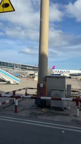 The video shows an airport tarmac on a cloudy day. In the foreground, there are several barriers and a concrete surface. To the left, a jet bridge labeled "Revolut" extends towards an airplane. The airplane, a Ryanair aircraft, is parked at the gate. Several airport vehicles, including a tug and a pushback tractor, are visible near the aircraft. In the background, other airplanes and airport buildings can be seen. The sky is blue with scattered clouds. The camera pans slightly to the right, focusing on the tail of the Ryanair plane, which features the company's logo.