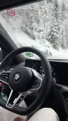 The video shows the interior view from the driver's seat of a BMW car driving on a snow-covered road. The driver's hands are on the steering wheel. The car's dashboard and digital display are visible, showing navigation information. Outside, snow-covered trees line the roadside. The car is in drive mode and moving at 68 km/h. A road sign with '210' is visible on the windshield.