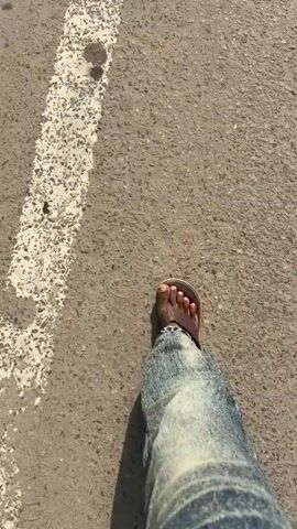 The video shows a first-person perspective of someone walking on an asphalt road. The person is wearing blue jeans and brown sandals, with their feet and lower legs visible. The road has a broken white line painted along its right side. In the background, there are some small dark objects on the road, possibly leaves or debris, and a small, dark, segmented insect. Further ahead, there is a concrete mixer truck parked on the side of the road, with a white and red striped barrier next to it. Overhead, a bridge structure, possibly for a metro line, is visible, supported by concrete pillars. Trees and vegetation are present on the left side of the road. The scene appears to be outdoors during the daytime, under a bright sky, with a generally dry environment.