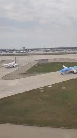 The video shows an aerial view of an airport tarmac on a cloudy day. An airplane is visible on the tarmac, appearing to be taxiing or preparing for takeoff. Several other airplanes and airport buildings are visible in the background, along with what appears to be a control tower. The tarmac is a light gray concrete with yellow and white markings. There are areas of green grass surrounding the tarmac. The camera seems to be positioned in another aircraft or a high vantage point.