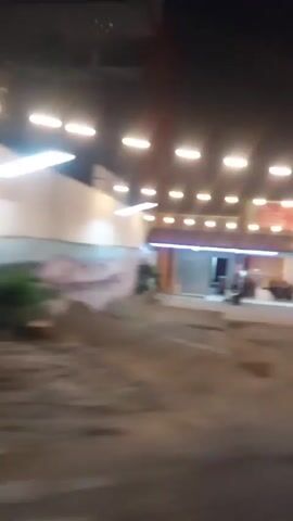 A blurry nighttime video shows a view from a moving vehicle. The vehicle passes by a brightly lit building with a long, curved facade and several windows or doors. The building has white columns and is illuminated by a row of lights along its roofline. Red and white flags are visible on a pole to the left. The ground is asphalt. The video is short and lacks clear details due to motion blur and low light conditions.