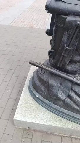 This media file showcases a bronze sculpture depicting a figure in historical attire, likely a soldier or explorer, based on the details of their gear. The sculpture is positioned outdoors on a paved plaza, indicated by the brickwork and a glimpse of a building in the background. The overall impression is of a monument commemorating a historical figure or event.

The camera focuses on the lower half of the sculpture, highlighting a large backpack or rucksack with rolled-up documents and straps. A rifle or musket is laid horizontally across the base of the sculpture, near the figure's feet. The detailed craftsmanship of the bronze is evident, with textures mimicking fabric, leather, and metal.

A hand, presumably belonging to the person filming, briefly interacts with the sculpture, touching the pouch on the backpack and then the base. This suggests a visitor engaging with the artwork.

The setting appears to be a public space, possibly a park or a square, with the paving stones and architectural elements of the building providing context. The lighting suggests it is daytime, but the exact time of day and weather conditions are not discernible. There is no visible text within the frame of the video. The emotion conveyed is one of historical reverence and appreciation for the artistry of the sculpture.