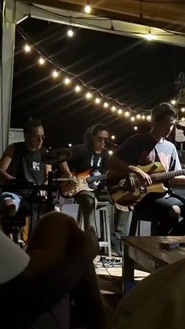 A band of three people performs live at an outdoor venue at night. The band members are all seated. The person on the right plays and sings into a microphone, playing an acoustic guitar. The middle person is playing an electric guitar, and the person on the left plays a keyboard and an electronic drumkit. The performance is under string lights, and the audience can be seen in the foreground. There is a wooden table in the right foreground.