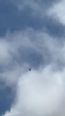 A helicopter is seen flying through a cloudy sky. The helicopter is small in the frame, appearing at a distance, and is primarily red and white. The sky is a mix of deep blue and white clouds, with the clouds appearing somewhat heavy and textured. The lighting suggests it is daytime. No people are visible in the media. The activity is simply the helicopter in flight against a backdrop of clouds. No text is visible in the media.