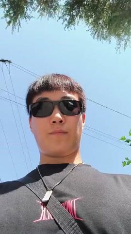 A close-up, low-angle shot of a young Asian man wearing sunglasses and a black t-shirt with a silver pendant necklace and a black strap across his chest. The man is looking directly at the camera with a neutral expression. The background shows a bright blue sky, green foliage at the top, and power lines with a utility pole on the left. The video appears to be taken outdoors on a sunny day.