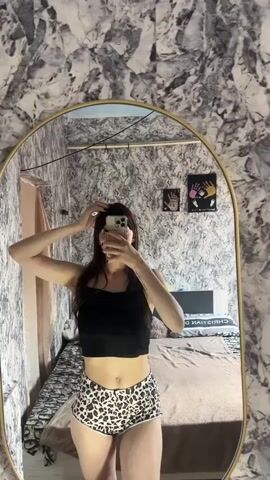 A young woman with dark hair is standing in front of a mirror, taking a selfie. She is wearing a black crop top and leopard print shorts. Her hair is pulled back from her face and she is touching it. The mirror is oval-shaped and has a gold frame. Behind her, a bed is visible, along with a patterned wallpaper on the wall. The room appears to be a bedroom.