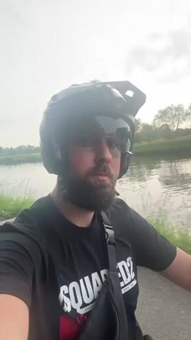 A man with a beard and wearing a black helmet, sunglasses, and a black t-shirt with "SQUARED2" printed on it, is riding a bike or motorcycle. The background shows a body of water, likely a river or canal, with greenery on the banks. The sky is overcast. The camera is presumably mounted on the handlebars, providing a first-person view of the man as he rides. The man looks directly at the camera.