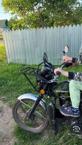 A man stands behind a child seated on a motorcycle. The child wears a camouflage shirt, light green pants with a pattern, and a gray helmet. The man wears a gray t-shirt and camouflage shorts. The motorcycle is black and green with a black rack on the front. They are outdoors in a grassy area next to a metal fence. The man's left hand is on the child's back, and his right hand is near the motorcycle's handlebars. The child's hands are on the handlebars. The man appears to be speaking to the child, who looks forward and then turns their head slightly to look at the man.