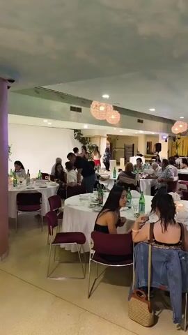 A group of people are gathered indoors at tables set for a meal. The room is lit and appears to be a restaurant or banquet hall, with decorative lights hanging from the ceiling. In the foreground, two women are seated at a table. Further back, a man in a black shirt and jeans is bending over to speak to someone at another table. Two women are standing near a white partition and appear to be dancing or gesturing. Several bottles of water and wine are visible on the tables.