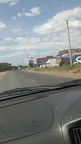 The video is shot from the dashboard of a vehicle driving on a paved road in a suburban area. There is a gas station, Rubis, on the right side of the road with a sign advertising the station's loyalty program. Many large bags are lined up on the side of the road, likely filled with some type of agricultural product. Several people are standing around these bags. A person is riding a motorbike on the right side of the road and there is light traffic in the opposite direction. The sky is blue with some scattered clouds. There are overhead power lines.