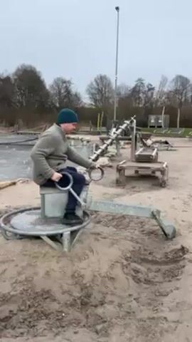 A man wearing a dark green beanie, a light gray sweater, dark pants, and dark shoes is sitting on a metal, playground-style excavator in a sandbox. He is operating the excavator's arm, moving it around in the sand. To the left of the sandbox is a shallow body of water. Behind the man are other playground structures and a light pole. A dog on a leash walks through the sandbox. In the background are bare trees and an overcast sky. In the audio, there is a child yelling, possibly crying, and the dog whimpering.