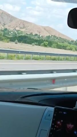 The video shows a view from inside a car as it travels on a highway. In the foreground, the dashboard of the car is visible, including a tablet or screen displaying an image of a person. The road stretches out ahead, with a white car passing by on the opposite lane. Beyond the road, there are hills with sparse vegetation and some buildings scattered on the slopes. The sky is partly cloudy. Reflective markers and a traffic sign with red and white stripes are present along the side of the road.