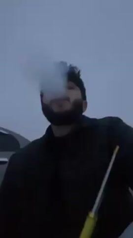 A low-angle shot shows a dark-haired man with a beard exhaling a large cloud of vapor into the overcast sky. He wears a dark shirt and holds a yellow and white smoking device. The sky is a uniform gray, and the side of a silver car is partially visible in the background. The man's expression is partially obscured by the vapor.