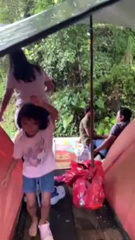 Within the frame, several individuals are gathered beneath a makeshift shelter, constructed with a tarp stretched over supporting poles. The setting appears to be outdoors, evidenced by the backdrop of lush greenery and trees visible beyond the shelter.

In the foreground, a young girl wearing a pink t-shirt and denim shorts stands prominently. She smiles and gestures with her arms, seemingly in a playful mood.

Behind the young girl, another woman, clad in a white shirt and dark shorts, is seen standing on a patterned mat. Her movements and gestures suggest she may be participating in some form of interaction or activity with other people in the scene.

Further back, seated on the mat beneath the shelter, are two men. They seem to be engaged in conversation or casual observation of the activities around them.

Scattered on the wooden floor of the shelter are various items, including bags, bottles, and other personal belongings. The scene depicts a casual, informal gathering of people enjoying an outdoor setting under a makeshift shelter, possibly during an event like a camping trip or picnic.