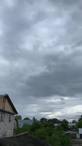 The video shows a view of a cloudy sky above a landscape of houses and trees. The sky is mostly covered with gray clouds, with a few patches of lighter areas. The houses in the background are small and surrounded by trees. The camera is static and there are no people visible in the frame. There are no actions taking place in the video.