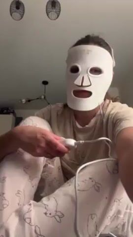 A woman in bunny-themed pajamas and a white LED face mask sits cross-legged, examining the mask's control panel. The panel is white and connected to the mask with a wire. The room has a modern chandelier and two hanging light fixtures. The woman’s fingernails are manicured.