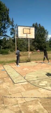 Three young men are playing basketball on an outdoor court with a worn surface. One of them is shooting the ball, while the other two are observing. The court is surrounded by trees and grass, and there is a building in the background. The sky is blue with some clouds. The basketball hoop is old and weathered.