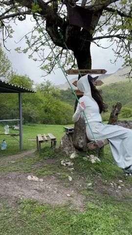 A woman in a white hoodie and light-colored pants is sitting on a wooden swing suspended from the branch of a large tree. She has dark hair and is wearing a baseball cap. She is gently swinging back and forth. Behind her is a grassy area with a small covered picnic area on the left. Two large water bottles sit on the picnic table. In the background, there is a view of green hills and a mountain range under an overcast sky. Attached to the tree trunk are signs pointing directions for "Horseriding," with phone numbers listed. The ground around the tree is a mix of dirt and grass. A bench is next to the tree. A male voice speaks to the woman on the swing.